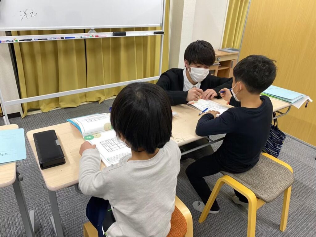2人の児童が講師と一緒に机で勉強している様子